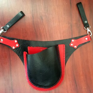 Pode incluir: Cinto de ferramentas de couro preto com acabamento vermelho e um único bolso. O cinto tem duas correias com clipes de metal.
