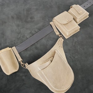 Pode incluir: Um cinto de couro bege com várias bolsas presas e uma bolsa grande. As bolsas são retangulares e quadradas, com costuras brancas e ferragens de metal. O cinto é marrom escuro.