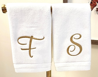 May include: Two white hand towels with gold embroidered letters 'F' and 'S' on a gold metal stand.