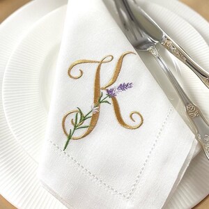 May include: A white linen napkin with a gold embroidered letter K and a small lavender flower design. The napkin is folded and placed on a white plate with a gold rim.