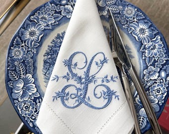 May include: A white linen napkin featuring a blue embroidered monogram of the letter L with floral accents. The napkin is folded and placed on a blue and white floral patterned ceramic plate alongside a silver fork and a silver knife.