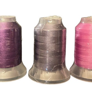 May include: Three spools of thread in different colors: purple, gray, and pink. The spools are in clear plastic containers.