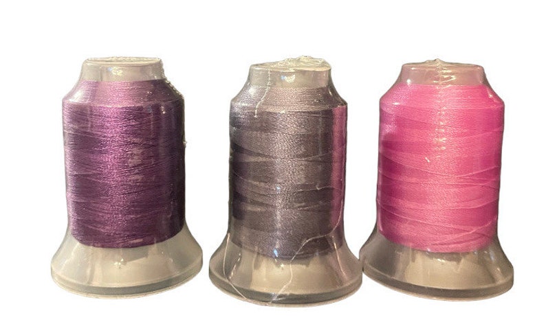May include: Three spools of thread in different colors: purple, gray, and pink. The spools are in clear plastic containers.