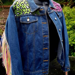 May include: A blue denim jacket with a unique design. The shoulders feature leopard print patches in green and pink. The left sleeve has colorful fringe. The jacket has a classic button-up front and two chest pockets.