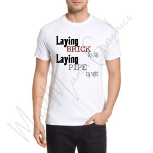 May include: White t-shirt with black text that reads "Laying Brick by day. Laying Pipe by night."