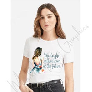May include: White t-shirt featuring a watercolour illustration of a woman in a blue dress. The text "She laughs without fear of the future." is printed on the shirt.