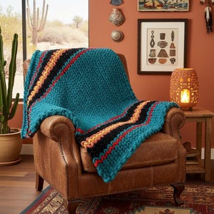 K&ouml;nnte beinhalten: Eine t&uuml;rkisfarbene Strickdecke mit schwarzen, roten und gelben Streifen, die &uuml;ber einem braunen Ledersessel drapiert ist. Die Textur der Decke ist grob und sichtbar. Der Raum hat ein s&uuml;dwestliches Thema mit einem Kaktus im Topf und Wanddekorationen.