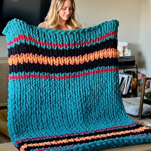 K&ouml;nnte beinhalten: Eine grob gestrickte Decke in T&uuml;rkis mit horizontalen Streifen in Schwarz, Rot und Orange. Die Decke besteht aus dickem, weichem Garn und bietet W&auml;rme und Komfort. Dieser dekorative &Uuml;berwurf ist ideal, um jedem Wohnraum Textur und Farbe zu verleihen. Ein gem&uuml;tliches Accessoire.