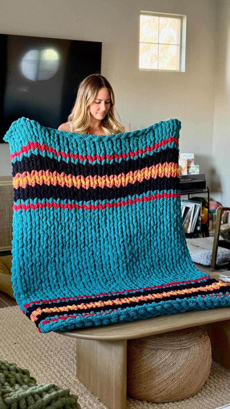 K&ouml;nnte beinhalten: Eine grob gestrickte Decke in T&uuml;rkis mit horizontalen Streifen in Schwarz, Rot und Orange. Die Decke besteht aus dickem, weichem Garn und bietet W&auml;rme und Komfort. Dieser dekorative &Uuml;berwurf ist ideal, um jedem Wohnraum Textur und Farbe zu verleihen. Ein gem&uuml;tliches Accessoire.