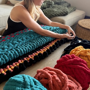K&ouml;nnte beinhalten: Eine Frau arbeitet an einer grob gestrickten Decke. Die Decke ist t&uuml;rkis, schwarz, rot und orange. Mehrere Kn&auml;uel Garn in T&uuml;rkis, Koralle, Rot und Gelb befinden sich im Vordergrund. Die Decke liegt auf einem Holzboden.