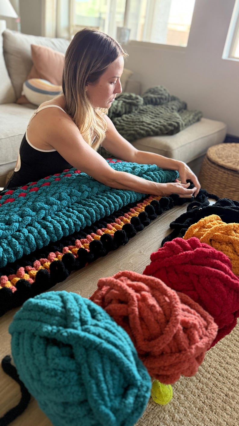 K&ouml;nnte beinhalten: Eine Frau arbeitet an einer grob gestrickten Decke. Die Decke ist t&uuml;rkis, schwarz, rot und orange. Mehrere Kn&auml;uel Garn in T&uuml;rkis, Koralle, Rot und Gelb befinden sich im Vordergrund. Die Decke liegt auf einem Holzboden.