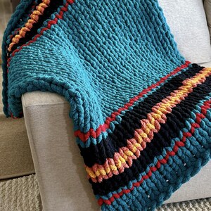 K&ouml;nnte beinhalten: Eine grob gestrickte Decke in T&uuml;rkisblau mit Streifen in Schwarz, Rot und Gelb. Die Decke ist &uuml;ber ein hellgraues Sofa drapiert und zeigt ihre Textur und Farbpalette. Das dicke Garn erzeugt einen gem&uuml;tlichen, einladenden Look.