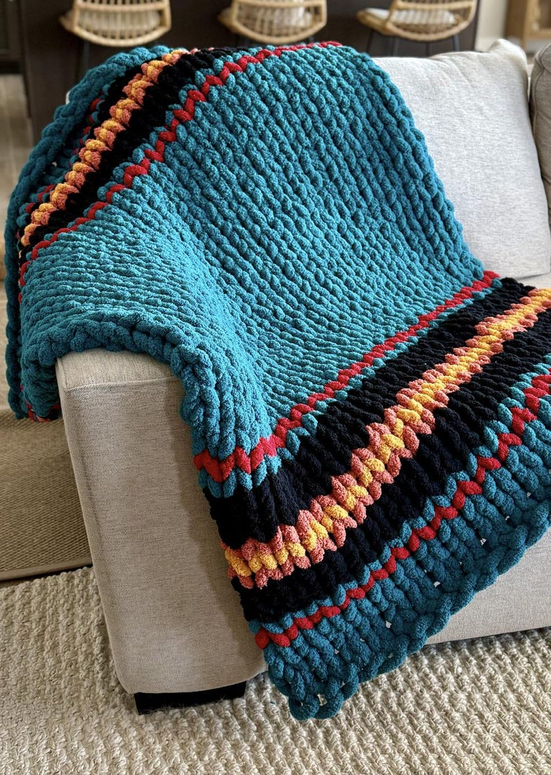 K&ouml;nnte beinhalten: Eine grob gestrickte Decke in T&uuml;rkisblau mit Streifen in Schwarz, Rot und Gelb. Die Decke ist &uuml;ber ein hellgraues Sofa drapiert und zeigt ihre Textur und Farbpalette. Das dicke Garn erzeugt einen gem&uuml;tlichen, einladenden Look.