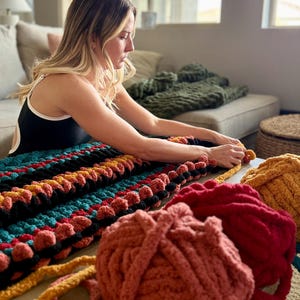 K&ouml;nnte beinhalten: Eine Frau arbeitet an einer gro&szlig;en, farbenfrohen, handgestrickten Decke. Die Decke hat Reihen in T&uuml;rkis, Schwarz, Orange und Gelb. Mehrere Kn&auml;uel Garn in Rot-, Orange- und Gelbt&ouml;nen liegen auf dem Tisch.