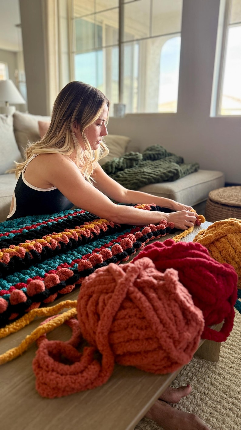 K&ouml;nnte beinhalten: Eine Frau arbeitet an einer gro&szlig;en, farbenfrohen, handgestrickten Decke. Die Decke hat Reihen in T&uuml;rkis, Schwarz, Orange und Gelb. Mehrere Kn&auml;uel Garn in Rot-, Orange- und Gelbt&ouml;nen liegen auf dem Tisch.