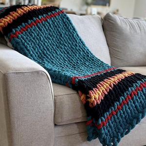 K&ouml;nnte beinhalten: Eine grob gestrickte Decke mit Streifen in T&uuml;rkis, Schwarz, Rot und Gelb, drapiert &uuml;ber einem grauen Sofa. Die Textur der Decke ist deutlich sichtbar und zeigt das dicke Garn und das handgestrickte Design. Die Farbpalette erzeugt eine gem&uuml;tliche Atmosph&auml;re.