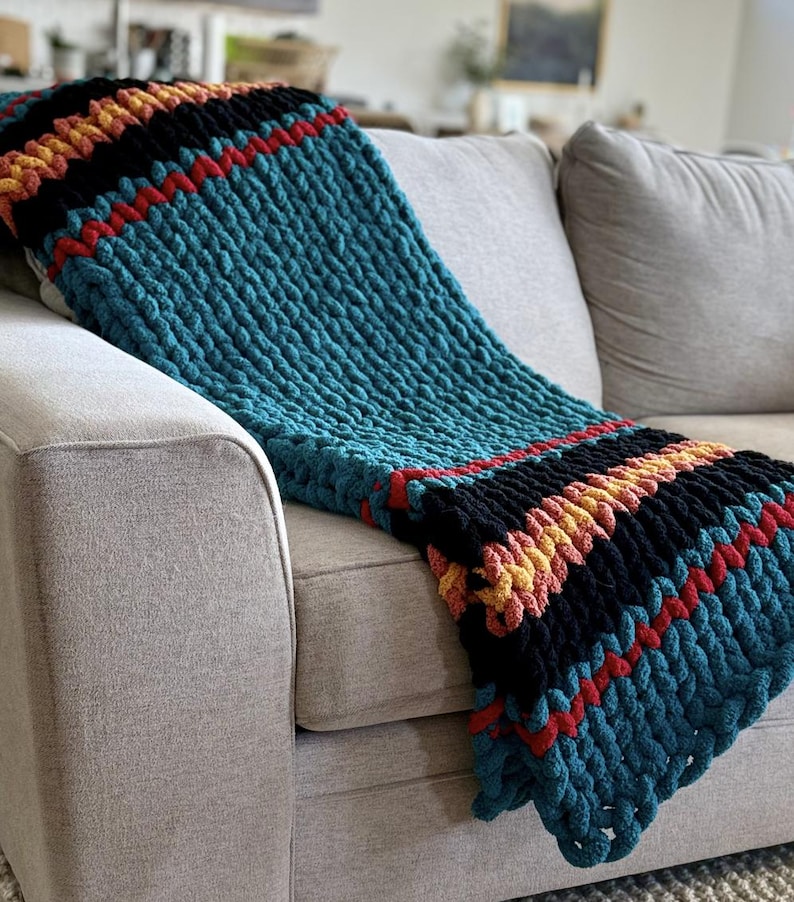 K&ouml;nnte beinhalten: Eine grob gestrickte Decke mit Streifen in T&uuml;rkis, Schwarz, Rot und Gelb, drapiert &uuml;ber einem grauen Sofa. Die Textur der Decke ist deutlich sichtbar und zeigt das dicke Garn und das handgestrickte Design. Die Farbpalette erzeugt eine gem&uuml;tliche Atmosph&auml;re.