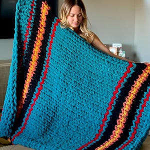 K&ouml;nnte beinhalten: Eine gro&szlig;e, t&uuml;rkisfarbene, grob gestrickte Decke mit diagonalen Streifen in Schwarz, Rot, Orange und Gelb. Die Decke wird pr&auml;sentiert und zeigt ihre Textur und Farbgebung. Perfekt f&uuml;r die Inneneinrichtung.