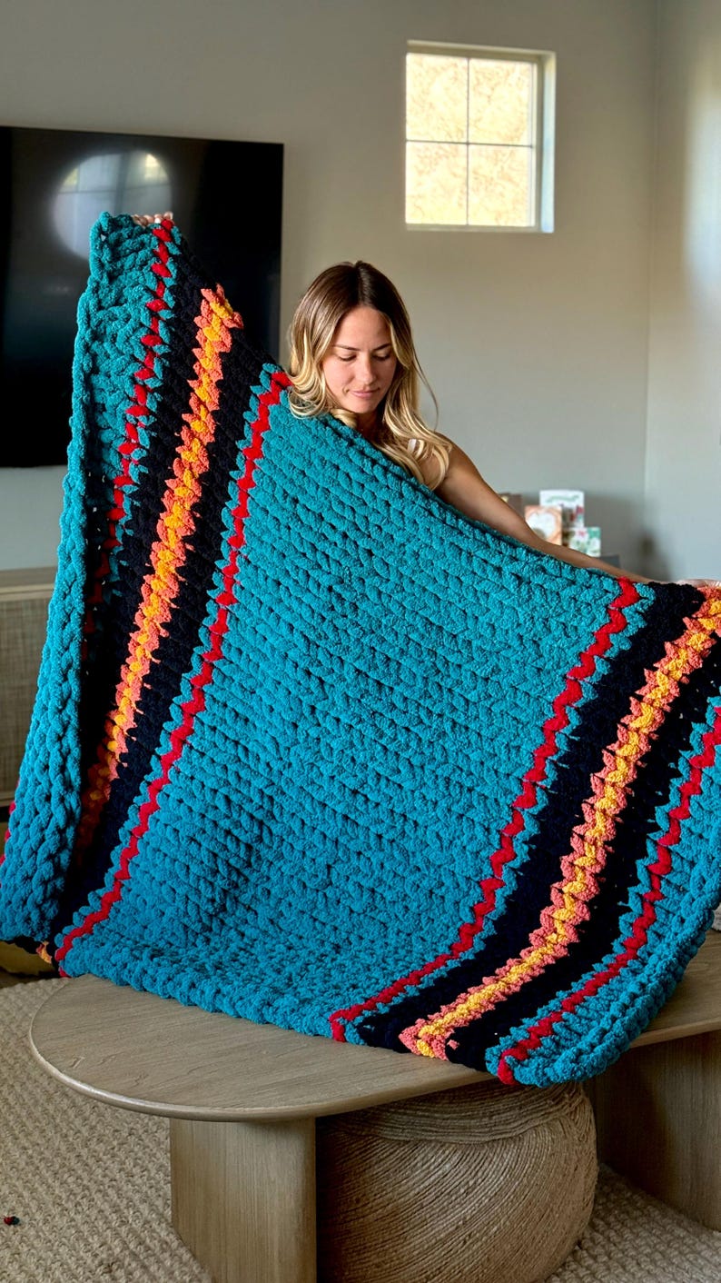 K&ouml;nnte beinhalten: Eine gro&szlig;e, t&uuml;rkisfarbene, grob gestrickte Decke mit diagonalen Streifen in Schwarz, Rot, Orange und Gelb. Die Decke wird pr&auml;sentiert und zeigt ihre Textur und Farbgebung. Perfekt f&uuml;r die Inneneinrichtung.