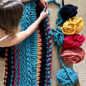 K&ouml;nnte beinhalten: Eine handgefertigte, grob gestrickte Decke in T&uuml;rkis, Schwarz, Rot und Gelb. Die Decke wird bearbeitet, mit verschiedenen farbigen Wollkn&auml;ueln in der N&auml;he. Eine Schere und Untersetzer liegen ebenfalls auf dem Tisch.