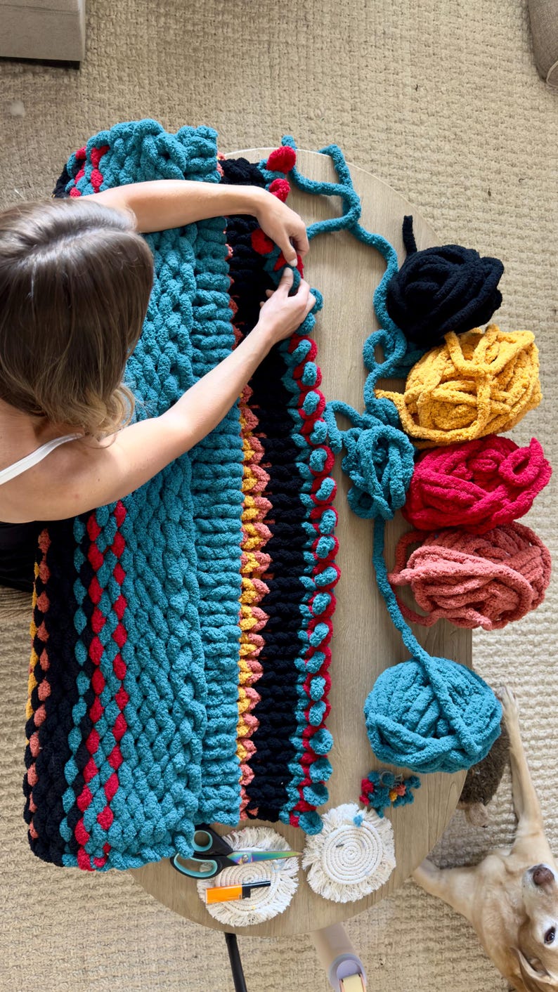 K&ouml;nnte beinhalten: Eine handgefertigte, grob gestrickte Decke in T&uuml;rkis, Schwarz, Rot und Gelb. Die Decke wird bearbeitet, mit verschiedenen farbigen Wollkn&auml;ueln in der N&auml;he. Eine Schere und Untersetzer liegen ebenfalls auf dem Tisch.
