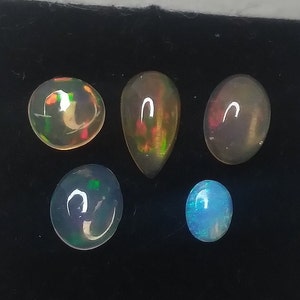 May include: Five opal gemstones of various shapes and colors, including round, teardrop, and oval, displayed on a black surface.