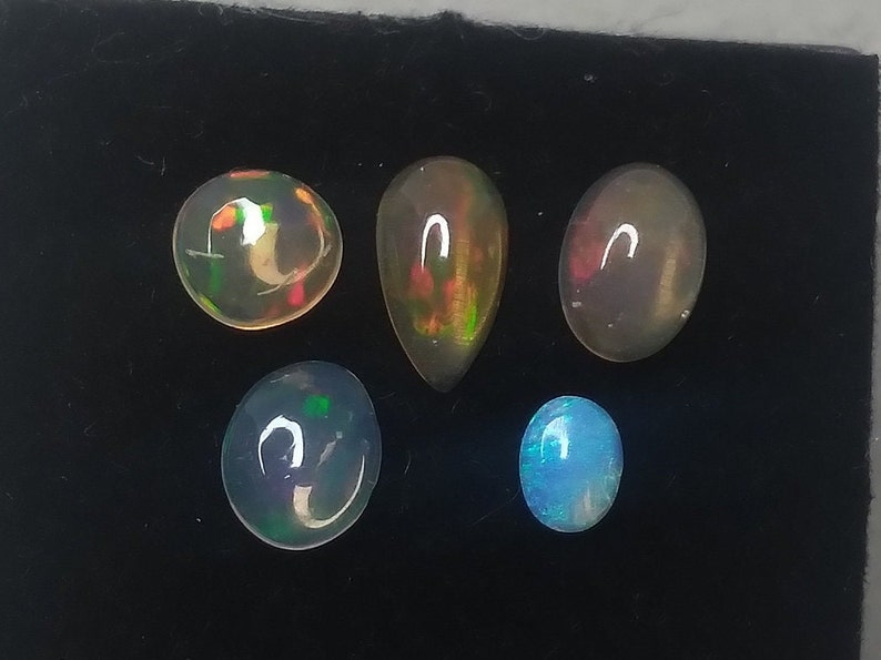 May include: Five opal gemstones of various shapes and colors, including round, teardrop, and oval, displayed on a black surface.