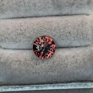 May include: A round, faceted gemstone with a reddish-brown hue. The gem is set against a gray, textured background, likely within a jewelry box or display case. The cut of the stone creates multiple facets that reflect light.