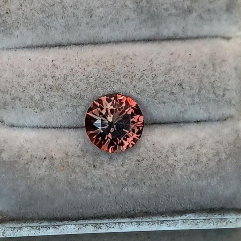 May include: A round, faceted gemstone with a reddish-brown hue. The gem is set against a gray, textured background, likely within a jewelry box or display case. The cut of the stone creates multiple facets that reflect light.