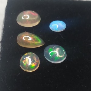 May include: Five opal gemstones of various shapes and sizes, including round, oval, and teardrop, displayed on a black velvet background. The opals exhibit a rainbow of colors, including green, blue, red, and orange.