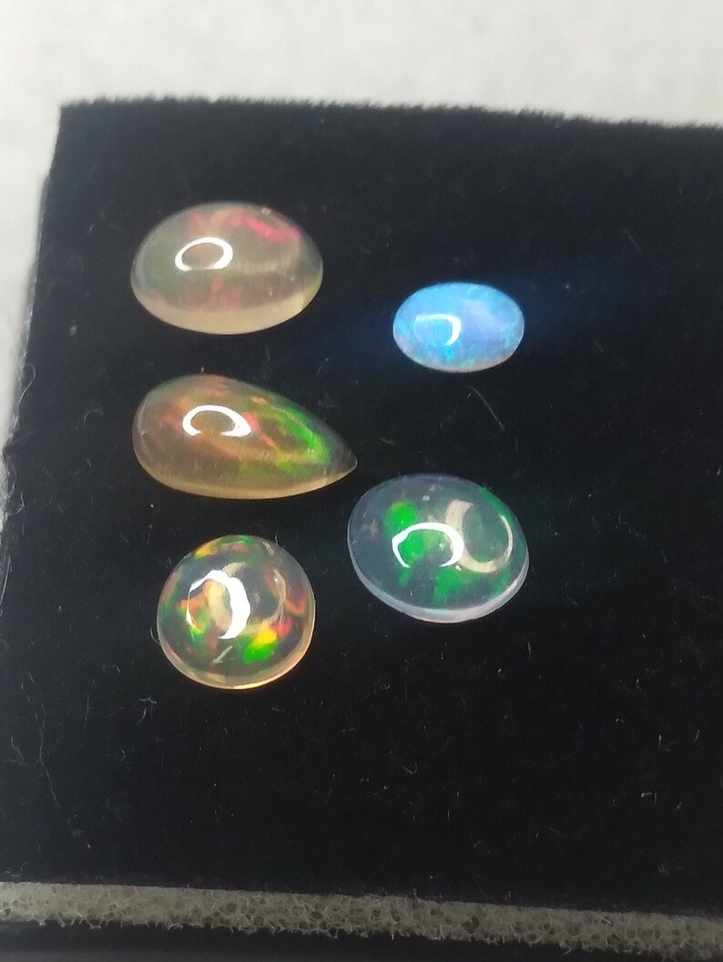 May include: Five opal gemstones of various shapes and sizes, including round, oval, and teardrop, displayed on a black velvet background. The opals exhibit a rainbow of colors, including green, blue, red, and orange.