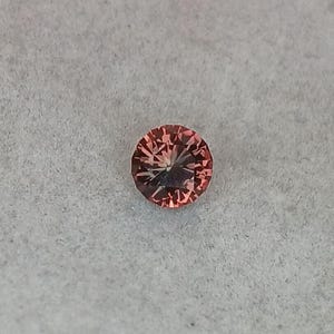 May include: A round, faceted gemstone with a peach-pink hue. The gem is set against a light gray background, showcasing its intricate internal structure and brilliance. The gemstone's facets catch the light, creating a sparkling effect.