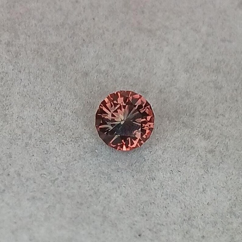 May include: A round, faceted gemstone with a peach-pink hue. The gem is set against a light gray background, showcasing its intricate internal structure and brilliance. The gemstone's facets catch the light, creating a sparkling effect.