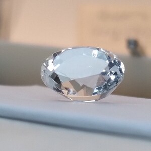 May include: A clear, faceted, oval-shaped crystal gemstone on a white surface.