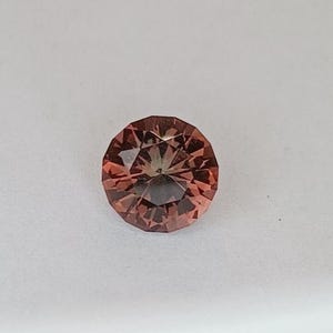 May include: A faceted, round gemstone with a reddish-brown hue. The gem has a complex internal structure, with multiple facets creating a play of light and shadow. The gemstone is set against a white background.