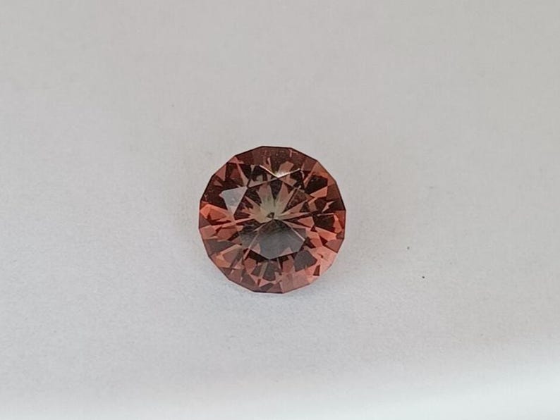 May include: A faceted, round gemstone with a reddish-brown hue. The gem has a complex internal structure, with multiple facets creating a play of light and shadow. The gemstone is set against a white background.