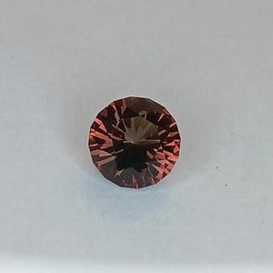 May include: A faceted, round gemstone with a reddish-brown hue. The gem has a dark center and radiating facets, creating a starburst effect. The gemstone is set against a white background.