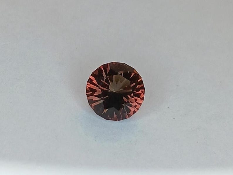 May include: A faceted, round gemstone with a reddish-brown hue. The gem has a dark center and radiating facets, creating a starburst effect. The gemstone is set against a white background.