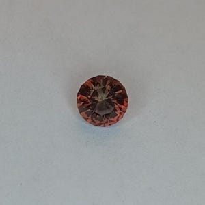 May include: A round, faceted gemstone with a reddish-orange hue. The gem is set against a white background, showcasing its intricate cut and color. The gemstone's facets reflect light, creating a sparkling effect.