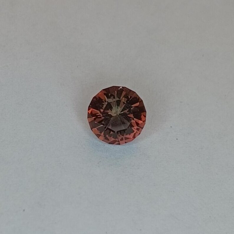 May include: A round, faceted gemstone with a reddish-orange hue. The gem is set against a white background, showcasing its intricate cut and color. The gemstone's facets reflect light, creating a sparkling effect.