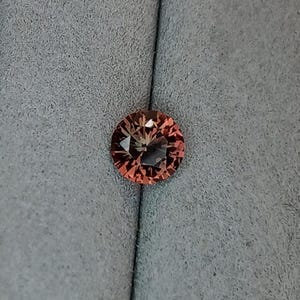 May include: A round, faceted gemstone with a peach and brown color gradient. The gem is set against a gray fabric background. The gemstone's cut creates a starburst effect, reflecting light.