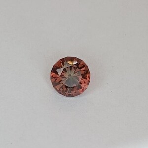 May include: A round, faceted gemstone with a warm, reddish-brown hue. The gem has a brilliant cut, reflecting light from multiple facets. The gemstone is set against a plain white background, highlighting its color and clarity. This gemstone is suitable for jewelry making.