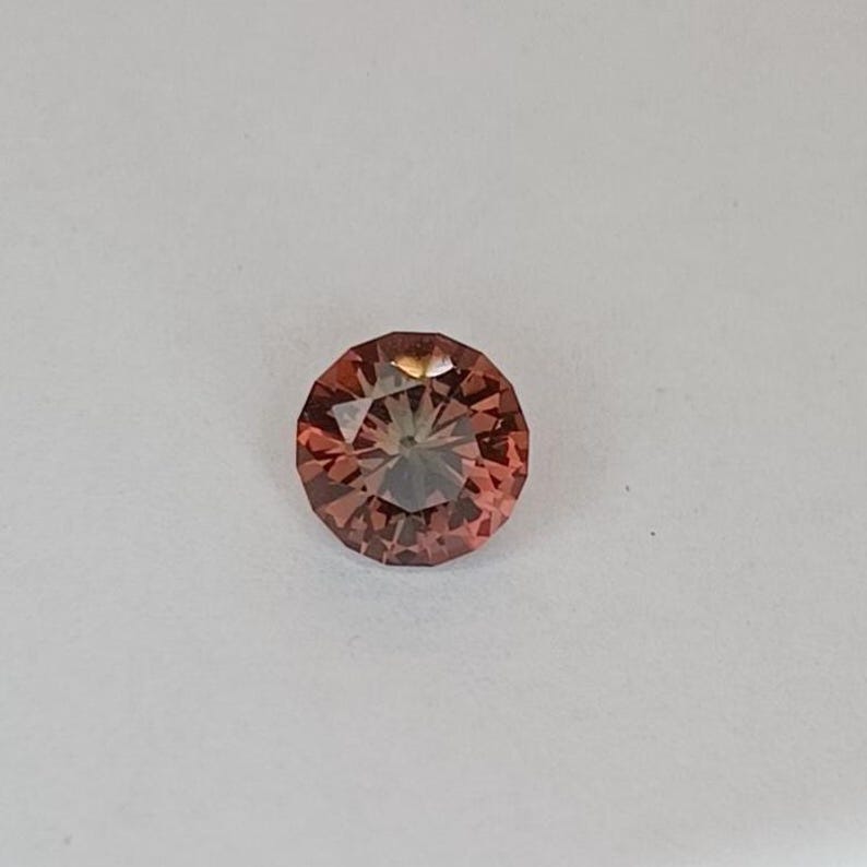 May include: A round, faceted gemstone with a warm, reddish-brown hue. The gem has a brilliant cut, reflecting light from multiple facets. The gemstone is set against a plain white background, highlighting its color and clarity. This gemstone is suitable for jewelry making.