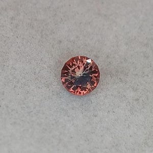 May include: A round, faceted gemstone with a peach-pink hue. The gem has a complex internal structure, with light reflecting off multiple facets. The gemstone is set against a light gray background.