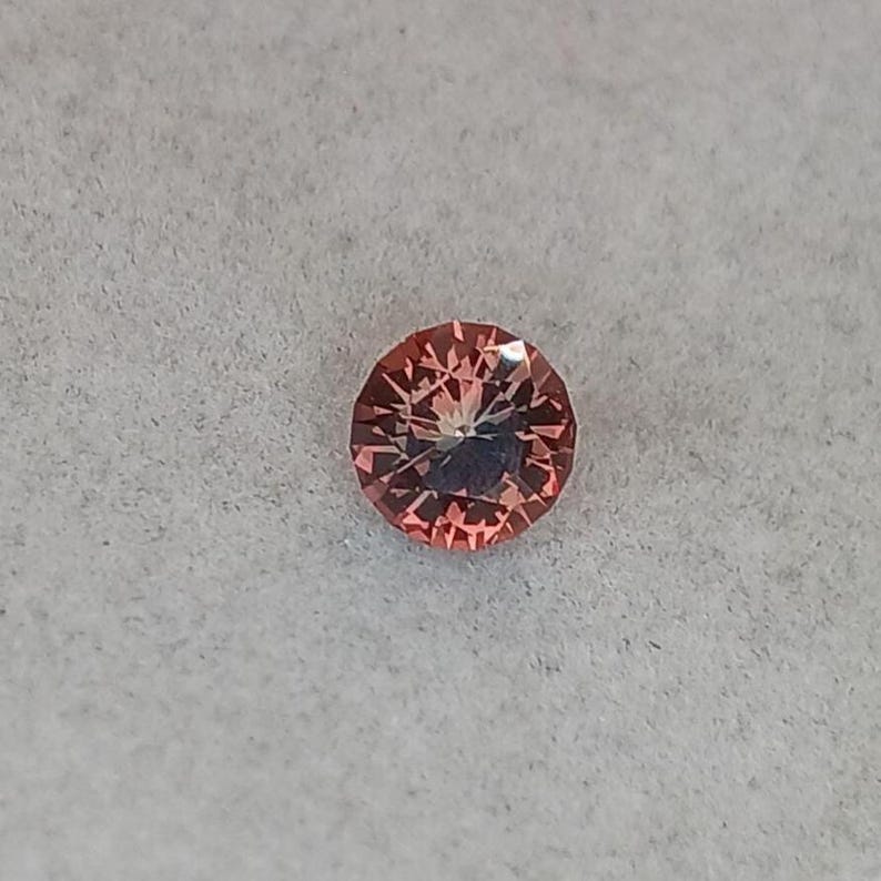 May include: A round, faceted gemstone with a peach-pink hue. The gem has a complex internal structure, with light reflecting off multiple facets. The gemstone is set against a light gray background.