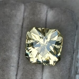May include: A light yellow gemstone with a square shape and a faceted cut. The gemstone is set in a clear, square setting.