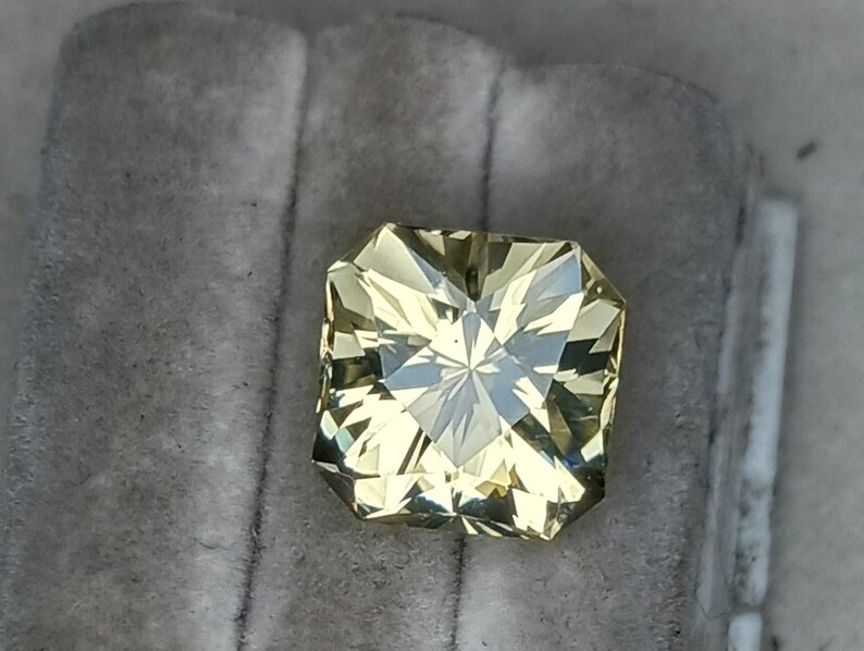 May include: A light yellow gemstone with a square shape and a faceted cut. The gemstone is set in a clear, square setting.