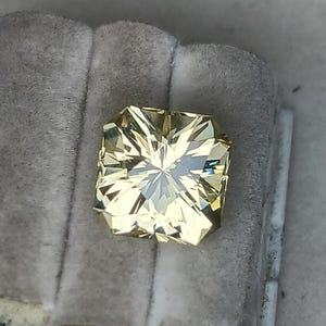 May include: A light yellow gemstone with a square shape and a faceted cut. The gemstone is set in a clear, square-shaped setting.