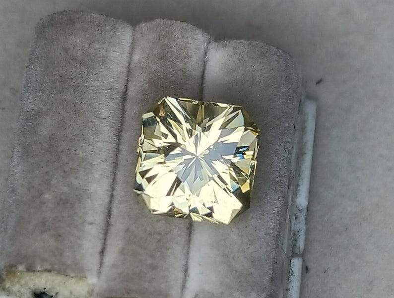 May include: A light yellow gemstone with a square shape and a faceted cut. The gemstone is set in a clear, square-shaped setting.