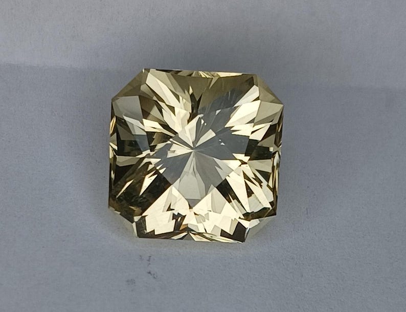 May include: A light yellow gemstone with a square shape and a faceted cut. The gemstone has a star-like pattern in the centre.
